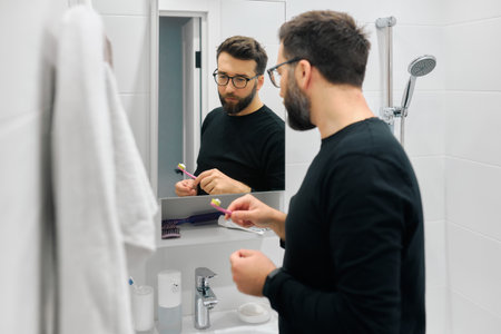 A man stands in a contemporary bathroom, focusing on his reflection while brushing his teeth. The morning light illuminates.の写真素材