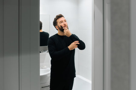 A man stands in a sleek bathroom, grooming his beard and adjusting his appearance in the mirror.の写真素材