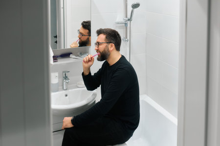 A man sits on the edge of a bathtub in a bright bathroom, brushing his teeth in preparation for the day.の写真素材