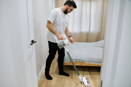 A man is cleaning the bedroom floor with a vacuum cleaner. The room features light walls and a neatly made bed.の写真素材