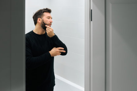 A man in a black long-sleeve shirt focuses on grooming his beard while standing in a well-lit bathroom.の写真素材