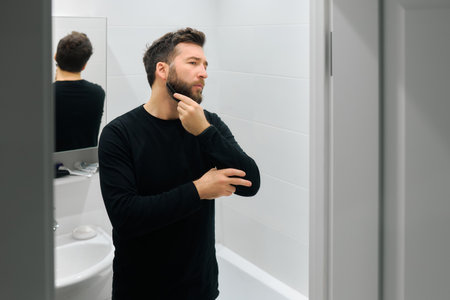 A man stands in front of a mirror in a bright bathroom, thoughtfully grooming his beard.の写真素材