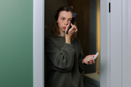 Woman stands in a bathroom, using a brush to apply makeup on her face. She is getting ready for the day.の写真素材
