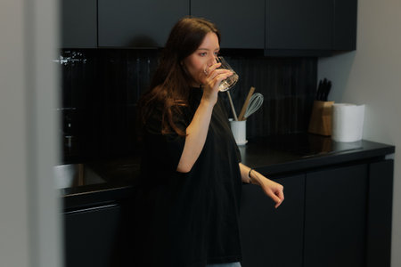 Woman stands in a black kitchen holding a glass of water. She appears focused and reflective as she takes a drink.の写真素材