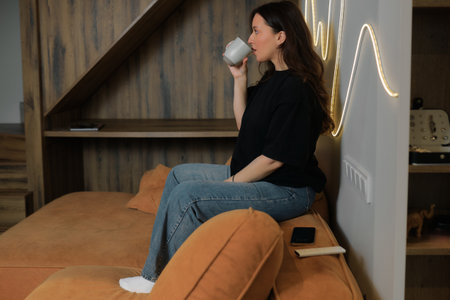 Woman in a black shirt sits on a couch with orange cushions. She holds a cup and gazes thoughtfully at the space around her.の写真素材