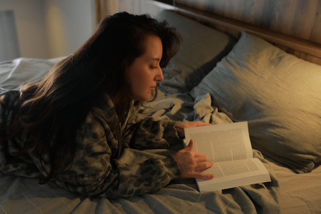 Person is lying on a bed, reading a book in a cozy room. The light from a lamp provides soft...