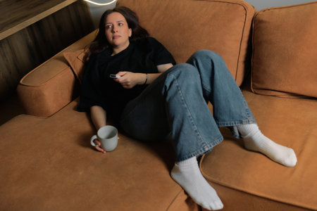 Woman relaxing on couch with remote and cup in cozy indoor spaceの写真素材