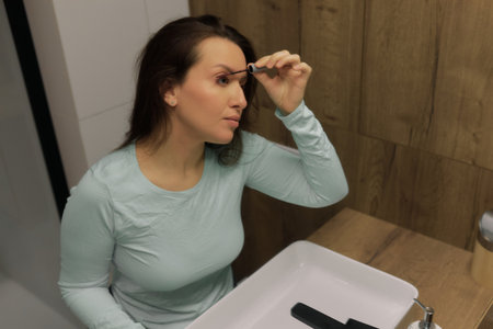 Woman sits at a bathroom sink applying mascara in front of a mirror. She is dressed casually.の写真素材