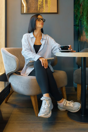 A young woman enjoys coffee while relaxing in a stylish cafe with modern decor in the afternoon sun, dressed casually.の写真素材