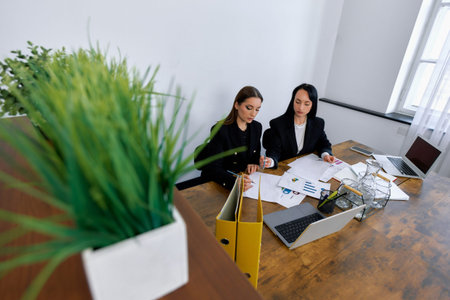 Two women are engaged in a business meeting at a modern office. They are reviewing documents and charts while using laptops. A potted plant adds a touch of greenery to the workspace.の写真素材