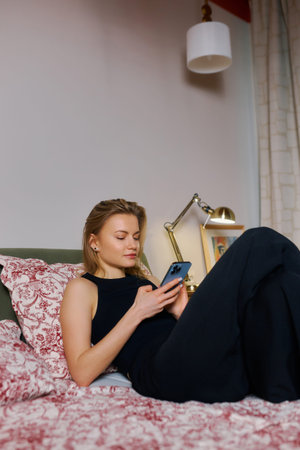 A young woman lounges comfortably on her bed, dressed casually in a black outfit. She is focused on...