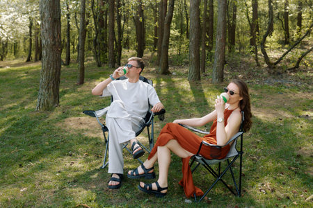 Two individuals sit in folding chairs surrounded by lush greenery, sipping from green cups. It is a...