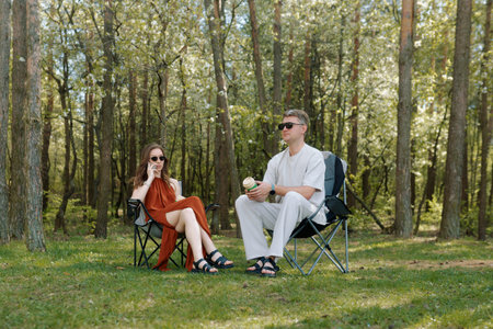 A couple is sitting on camping chairs in a serene forest setting. They are enjoying drinks while basking in the warm sunlight filtering through the trees.の写真素材