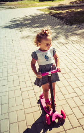 Cute child skilfully skates on a pink two-wheeled scooter on a summer sunny dayの写真素材
