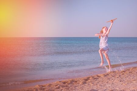 A cheerful girl plays with her toy airplane and jumps on the waves of a sandy beach in bright sunlight.の写真素材