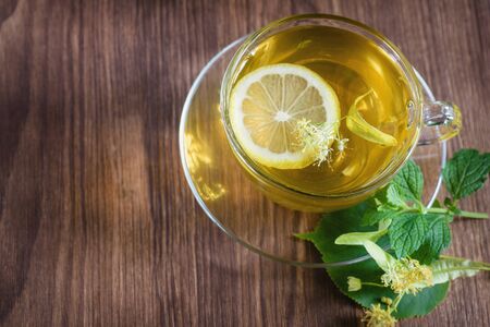 A cup of medicinal, lime tea, standing on a wooden table, in the rays of sunlight, there are flowers of chamomile lime trees around.の写真素材