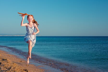 A happy red-haired child, playing with a toy airplane, runs along the sandy beach of the sea against the blue summer sky.の写真素材