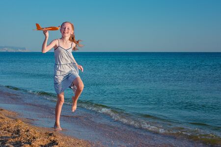 Beautiful red-haired girl, playing with a toy airplane, runs along the beach of the ocean against the blue summer sky and enjoys the rays of the sun and freedom.の写真素材