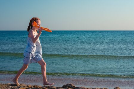 A beautiful girl, playing with a toy airplane, runs along the sandy beach of the sea against the background of a clear blue sky and enjoys the rays of the summer sun.の写真素材