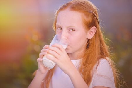Red-haired girl drinks fresh milk from a glass on the background of a beautiful sunny morning in the village.の写真素材