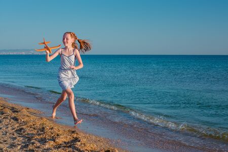 Red-haired girl, playing with a toy airplane, runs along the beach of the sea against the blue summer sky and enjoys the rays of the sun and freedom.の写真素材