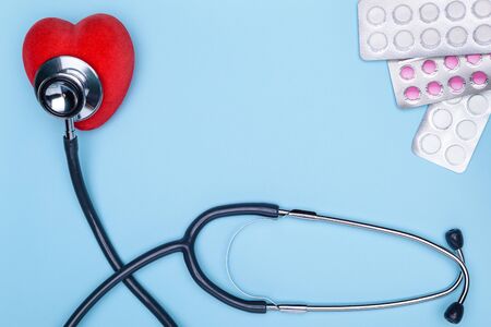 Blue background lies a statoscope, a red saddle and pills. The concept of treatment of cardiovascular diseases. Closeup of painted heart, lot of drugs and stethoscope on blue background.の写真素材