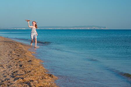 Happy red-haired girl, playing with a toy airplane, runs along the sandy beach of the sea against the blue summer sky and rejoices the sun.の写真素材