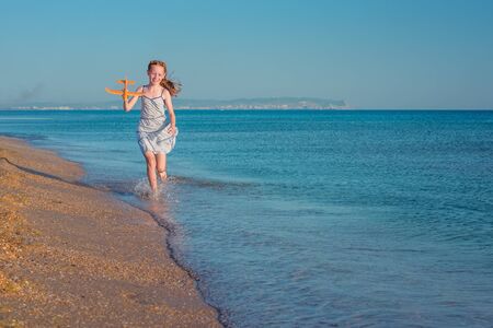 Cheerful red-haired girl playing with toy airplane and jumps over the waves of the sandy shore of the sea against the background blue summer sky and rejoices in the rays of the sun and freedom.の写真素材