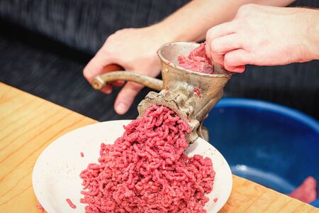 Chef prepares minced meat from fresh meat, background for the store. Minced meat. Woman's hands twist manual meat grinder through which minced meat comes out. Minced meat in minced meat is passed through a manual meat grinder close-up. の写真素材