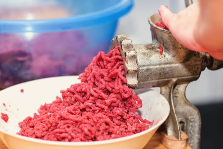  Woman's hands twist manual meat grinder through which minced meat comes out. Chef prepares minced meat from fresh meat, background for the store. Minced meat. Minced meat in minced meat is passed through a manual meat grinder close-up. の写真素材