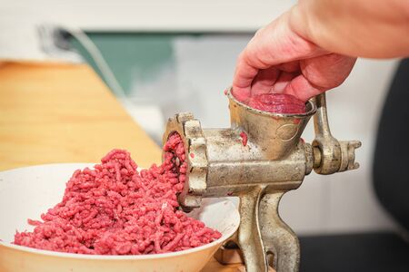 Minced meat. Woman's hands twist manual meat grinder through which minced meat comes out. Minced meat in minced meat is passed through a manual meat grinder close-up. Chef prepares minced meat from fresh meat, background for the store.の写真素材