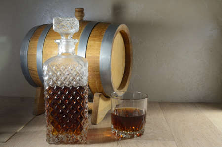 An oak barrel on a stand, a glass decanter and a glass on the table against the background of the...