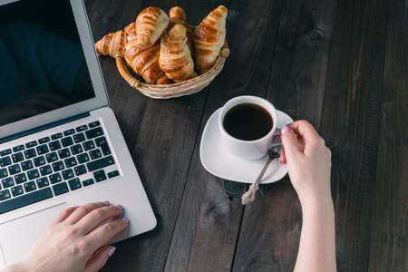 Basket with tasty fresh croissants and hot morning cup of coffee or tea with female working at laptop computer in background.の写真素材