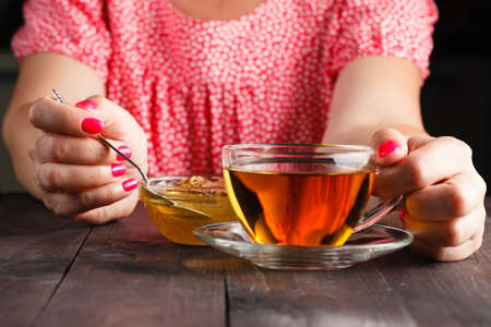 Woman hold cup of hot tea in glass cup, jar of honey, honey dipper on the old wooden backgroundの写真素材