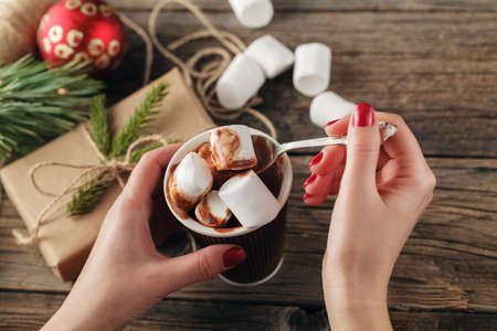 female hand holding cup of hot cocoa or chocolate with marshmallow on wooden table from aboveの写真素材