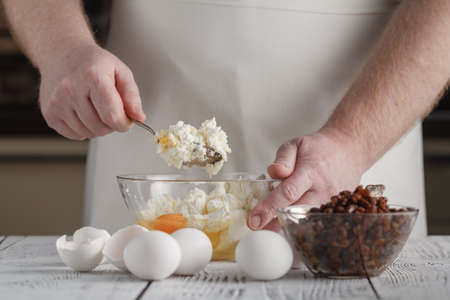 man's hands mixing cottage cheese, eggs, flour, raisins in the bowl on the table in the kitchen. Tasty, sweet meal. Healthy eating and lifestyle.の写真素材