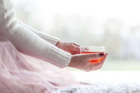 Woman in a grey sweater and warm socks holding a cup of tea while sitting on a white knitted blanketの写真素材