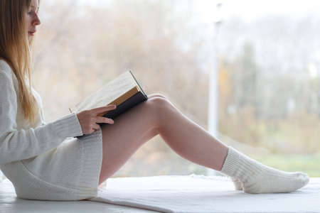 Soft cozy photo of slim tan woman in warm sweater and woolen socks with book in handsの写真素材