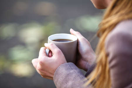 Closeup view of disposable cup of coffee or tea in womans hand. Autumn park with trees and yellow leafesの写真素材