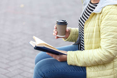 woman sitting on bench and reading book while drinking takeaway coffee from paper cup