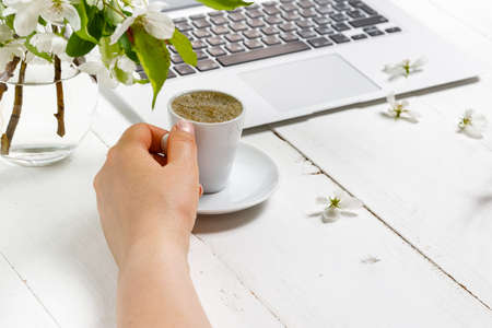 Feminine business concept with laptop, spring flowers bouquet and woman's hands holding cup of warm tea or coffeeの写真素材
