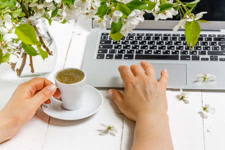 Feminine business concept with laptop, spring flowers bouquet and woman's hands holding cup of warm tea or coffeeの写真素材