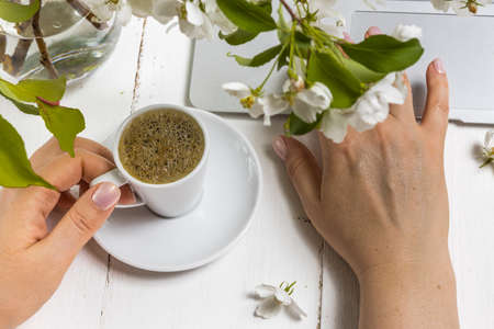 Workspace with girl's hand on laptop keyboard and cup of coffee, white spring apple tree flowers on white woodden background. View feminine office table desk. Freelancer working placeの写真素材