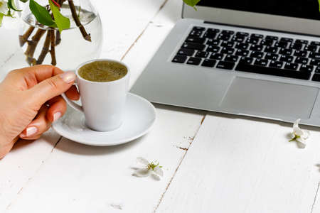 Feminine business concept with laptop, spring flowers bouquet and woman's hands holding cup of warm tea or coffeeの写真素材