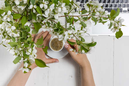 Feminine business concept with laptop, spring flowers bouquet and woman's hands holding cup of warm tea or coffeeの写真素材