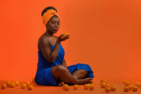Beautiful african woman with vivid makeup. Beauty portrait of sensual model with colorful makeup on orange background with fruitsの写真素材