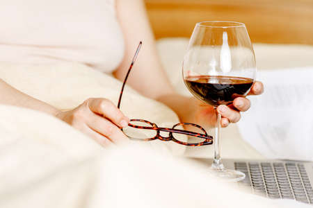 woman working on a laptop laying on the bed in the house. Freelancer woman working on her laptop at home with glass of wineの写真素材