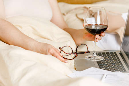 woman working on a laptop laying on the bed in the house. Freelancer woman working on her laptop at home with glass of wineの写真素材
