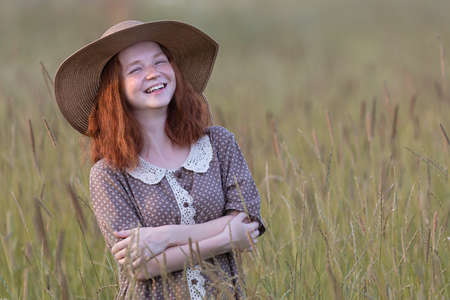 emotional redhead girl stands in the grass on a summer meadow at sunset in an elegant dressの写真素材