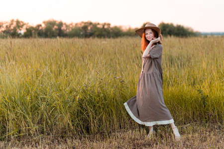 redhead girl posing on a summer meadow at sunset in an elegant dressの写真素材